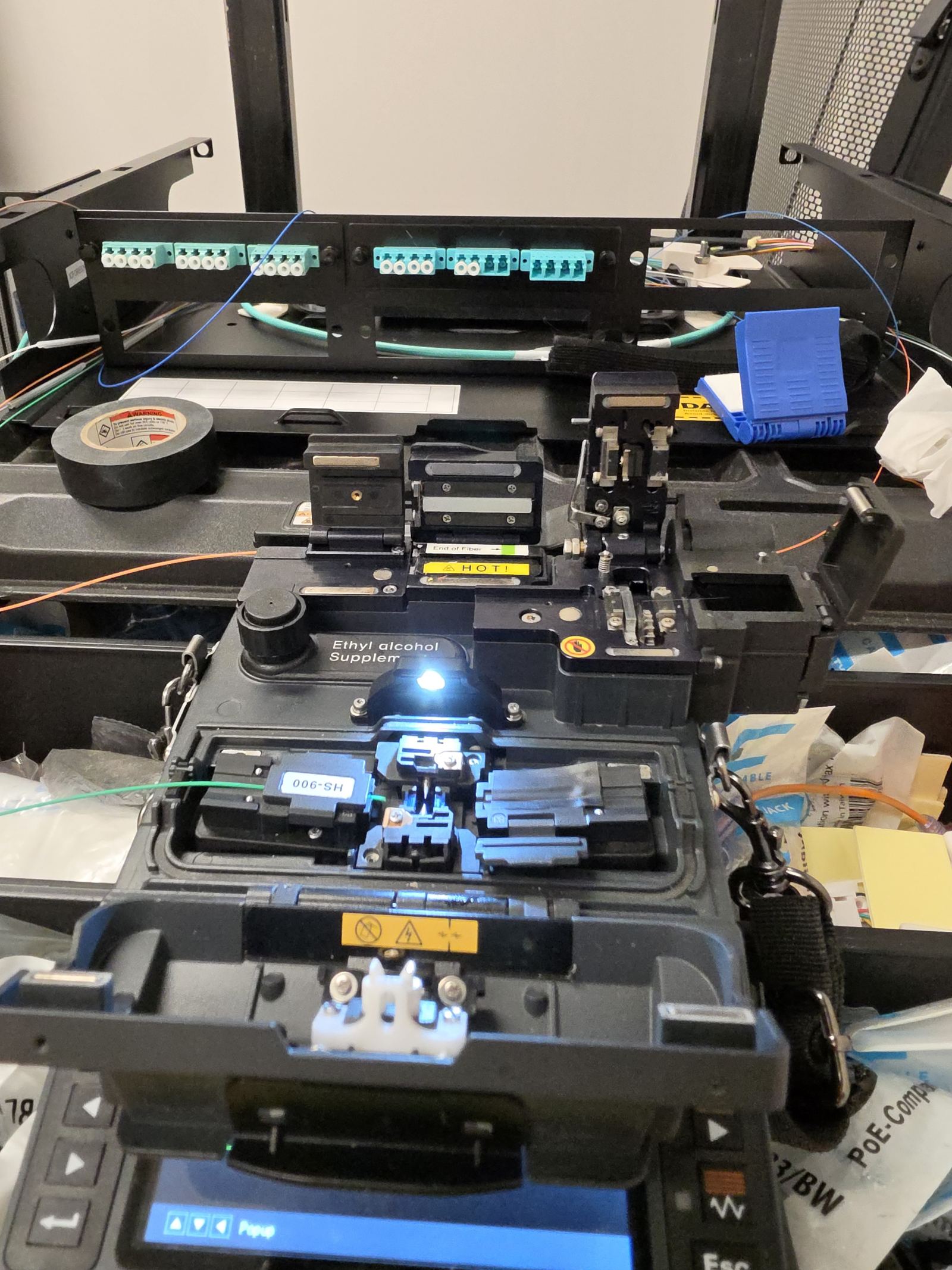 Fusion splicer mid-splice in front of rack-mounted LC fiber patch panels with aqua adapters in a large warehouse in Mount Pleasant, TN